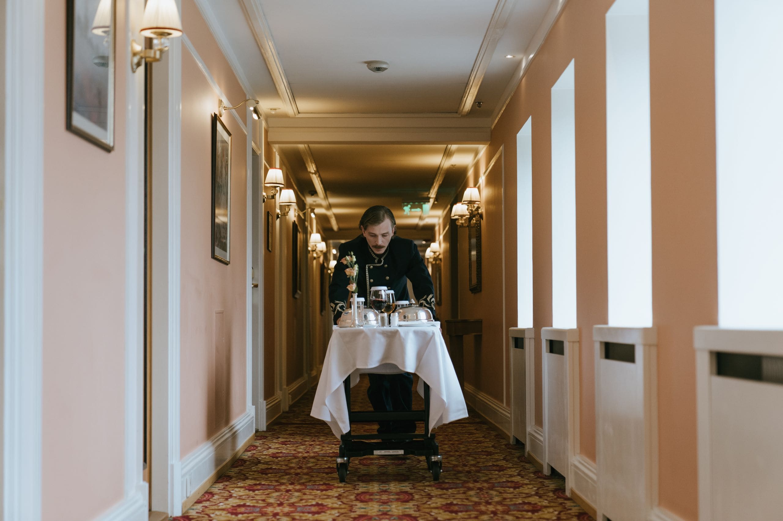 A host greeting an arriving guest in the lamplit entrance hall of a private house