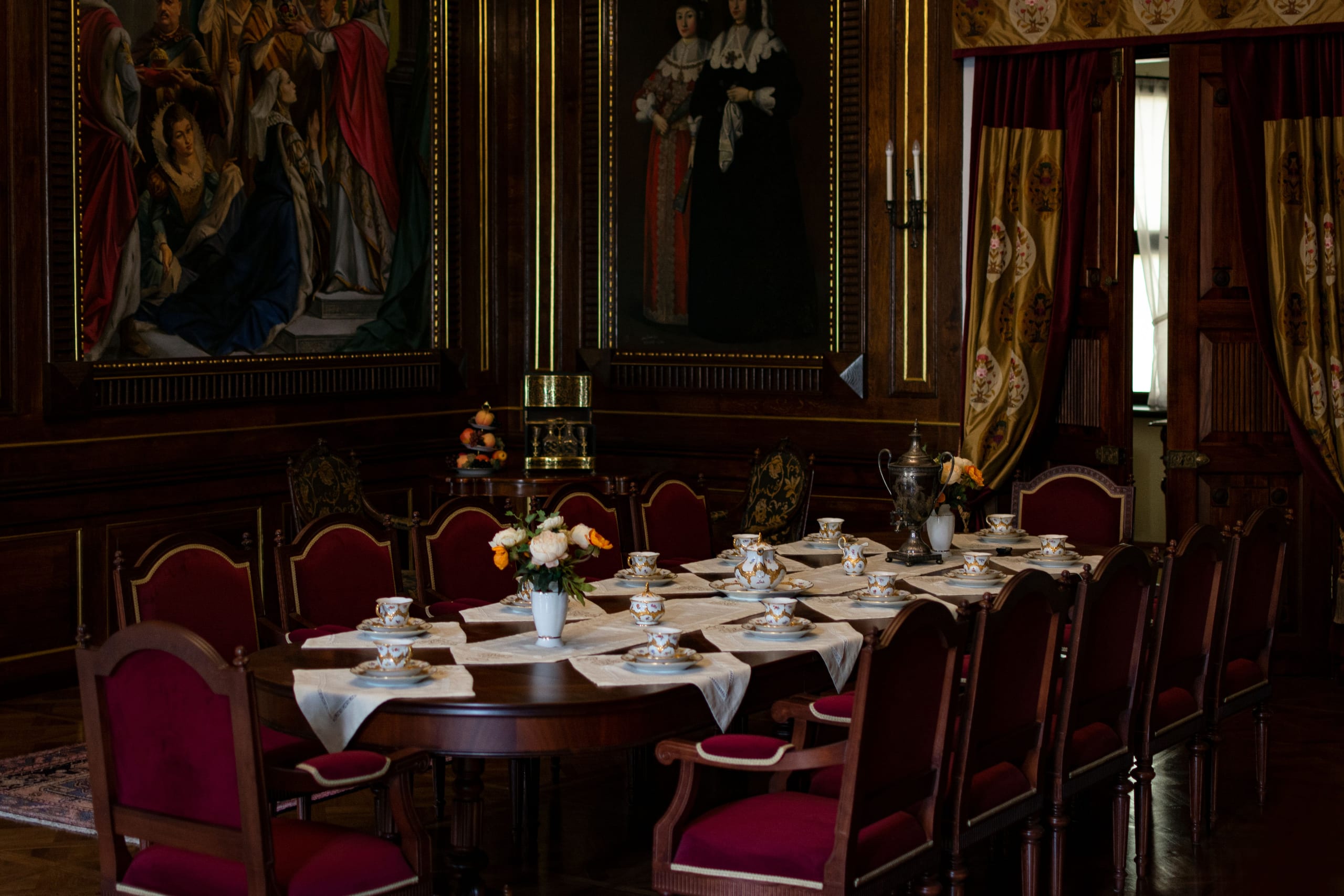 An established dining room hung with oil paintings, its long table laid with china between rows of crimson chairs