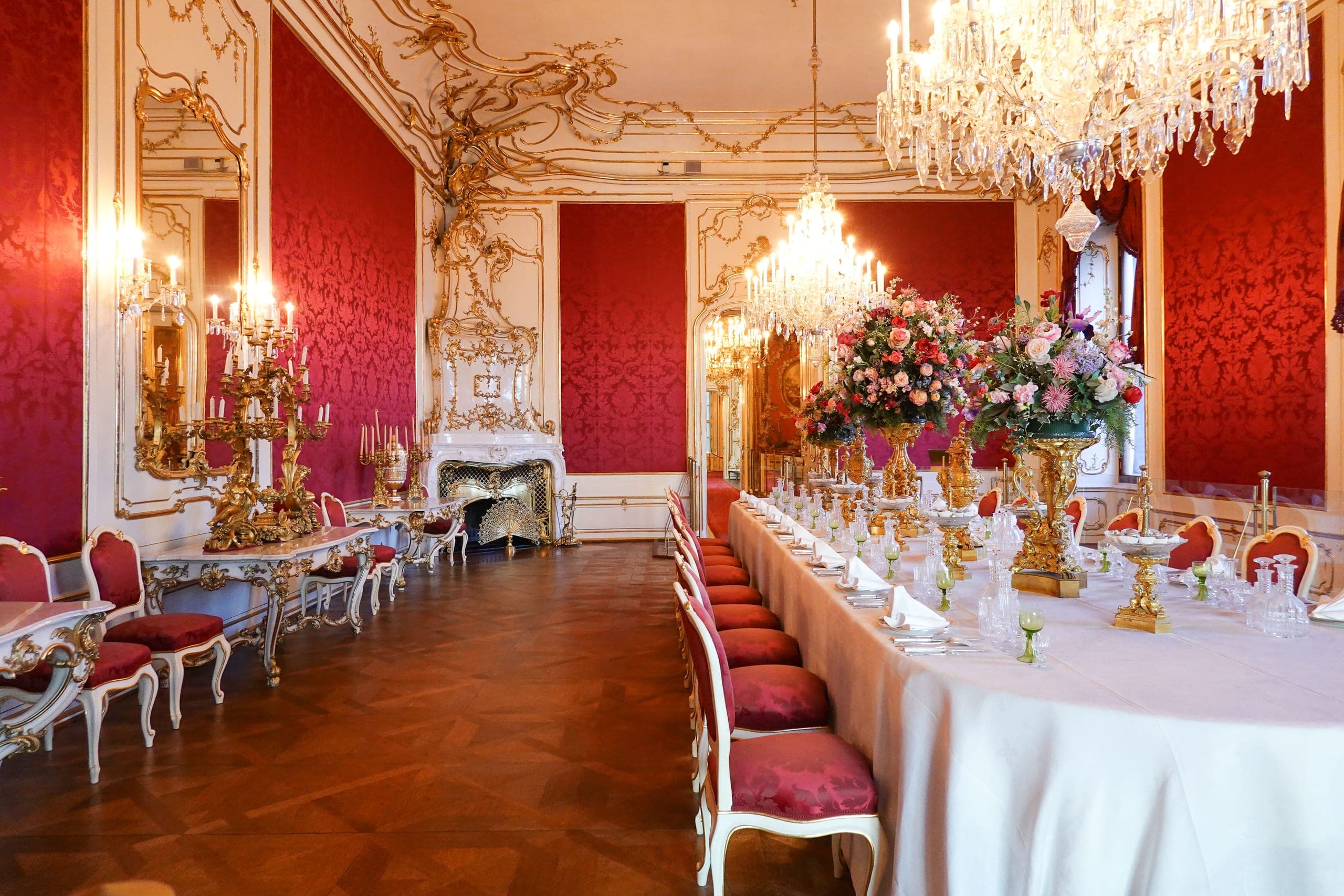 A grand crimson and gilt banquet room, its long table dressed in white and set with gold candelabra and flowers